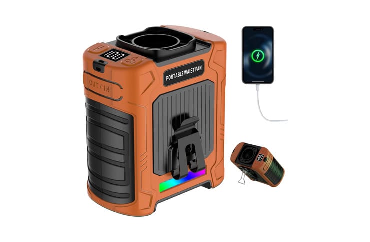 Orange portable waist fan power unit with a built-in fan, digital display, and USB charging a smartphone nearby. A small accessory device is placed beside it.